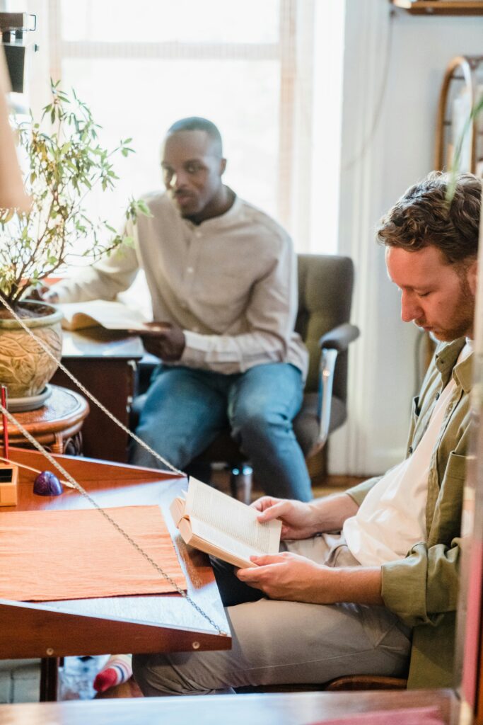 Two men enjoy reading books in a cozy, sunlit apartment setting, fostering relaxation.